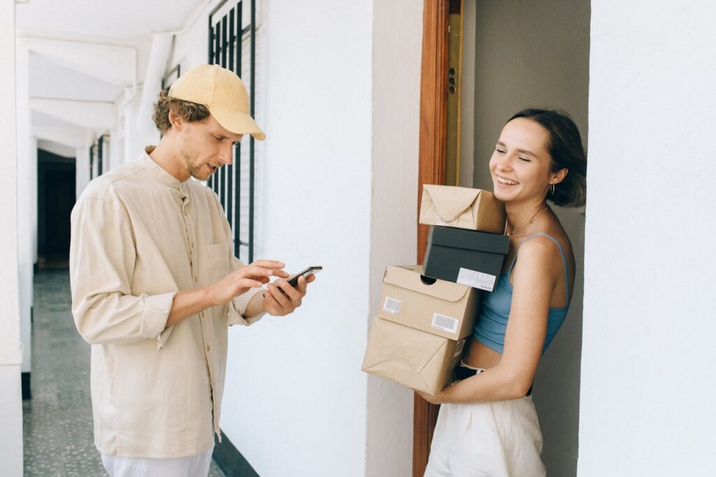 A cheerful woman receives a delivery from a person in a hallway, using a smartphone for online payment.